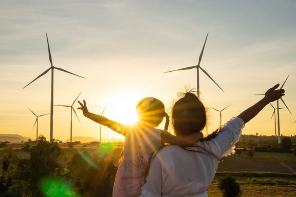 Eine Frau steht mit einem kleinen Mädchen auf dem Arm im Sonnenuntergang vor einem Windpark.